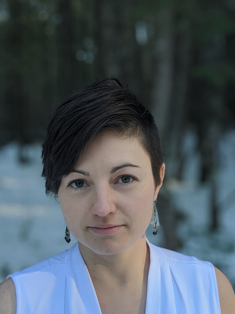 Portrait of Sue Ishaq. She is wearing a white sleeveless blouse and silver earrings. She has short, strait brown hair and is looking directly at the came. She is standing in a snowy forest. 