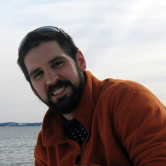 Portrait of Dr. Kiley Daley. He is wearing an orange fleece jacket and sunglasses on his head. He has short brown hair and a beard. He is smiling at the camera and is outdoors in front of a lake or the ocean.