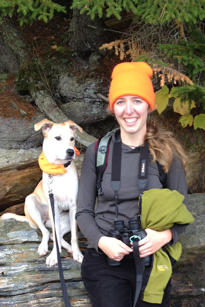 Alice Doughty rests on a log during a hike with her dog. She is reaching binoculars around her neck. 