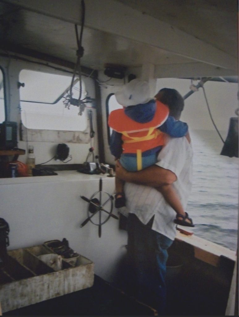 A man stands at a steering wheel while holding a toddler in an orange pfd inside the cabin of a lobster boat.