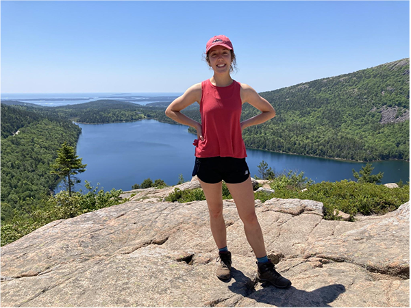 Roadcap stands on a rock that overlooks a lake surrounded by forests. The ocean and sky is in the horizon.