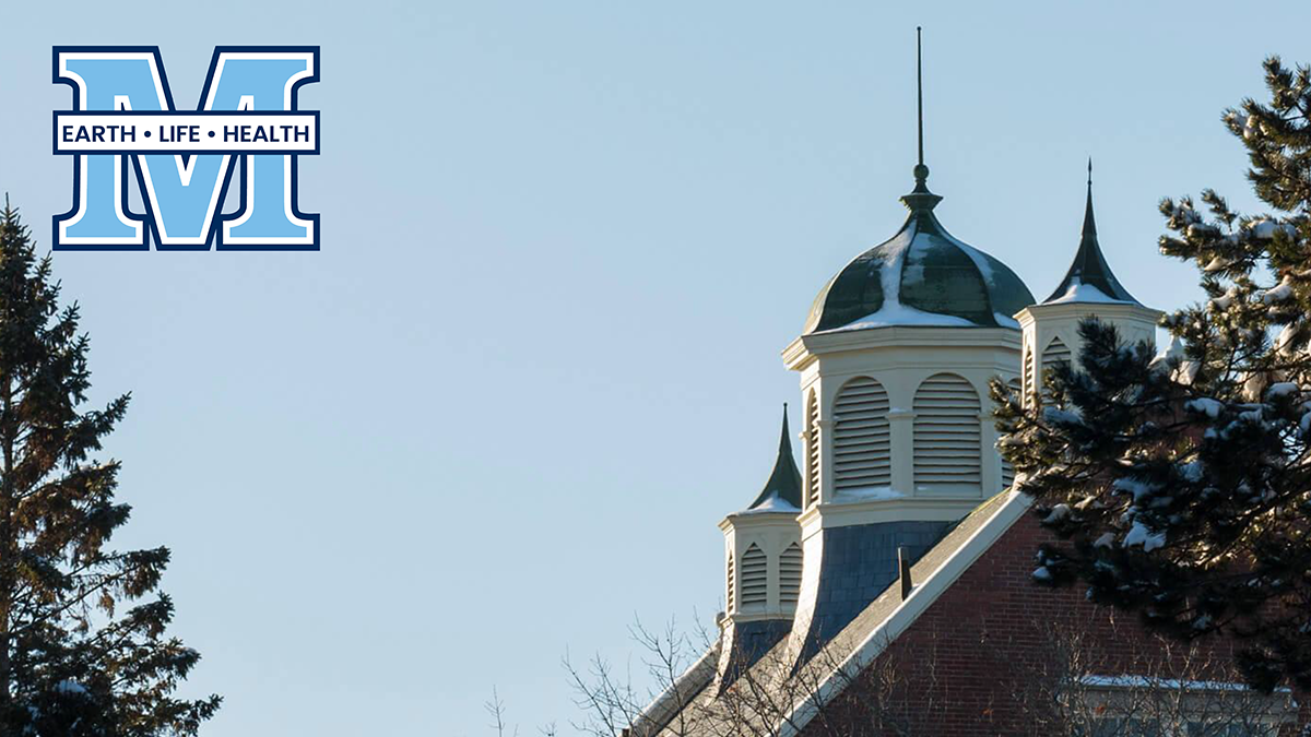 A photo of Winslow Hall's roofline in the winter with UMaine's M logo featuring the words "earth life health"